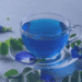 A clear glass teacup filled with bright blue butterfly pea flower tea, sitting on a glass saucer. Fresh green vines and blue butterfly pea flowers are arranged around the cup on a grey background. This image is featured on MyDailyDispatch.