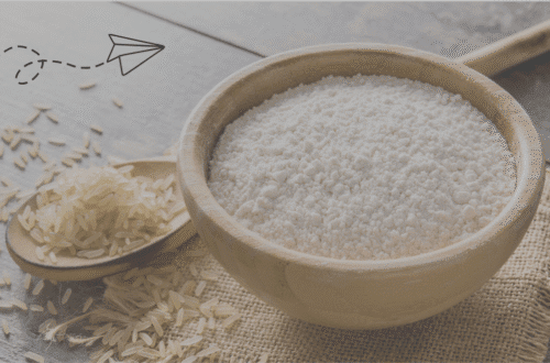 A wooden bowl overflowing with white rice flour next to a wooden spoon holding raw, unpolished rice grains, set on a rustic wooden surface with a burlap accent. The image highlights ingredients for a homemade rice flour face pack, a natural solution for glowing skin.